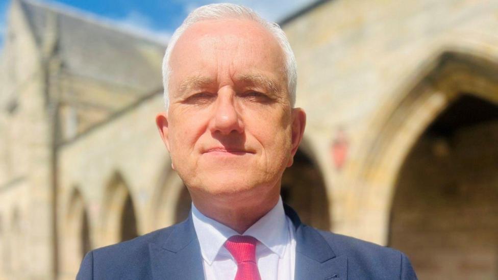 Prof Peter Edwards, a man with white hair and wearing a blue suit jacket, light shirt and red/pink tie, in a university setting.