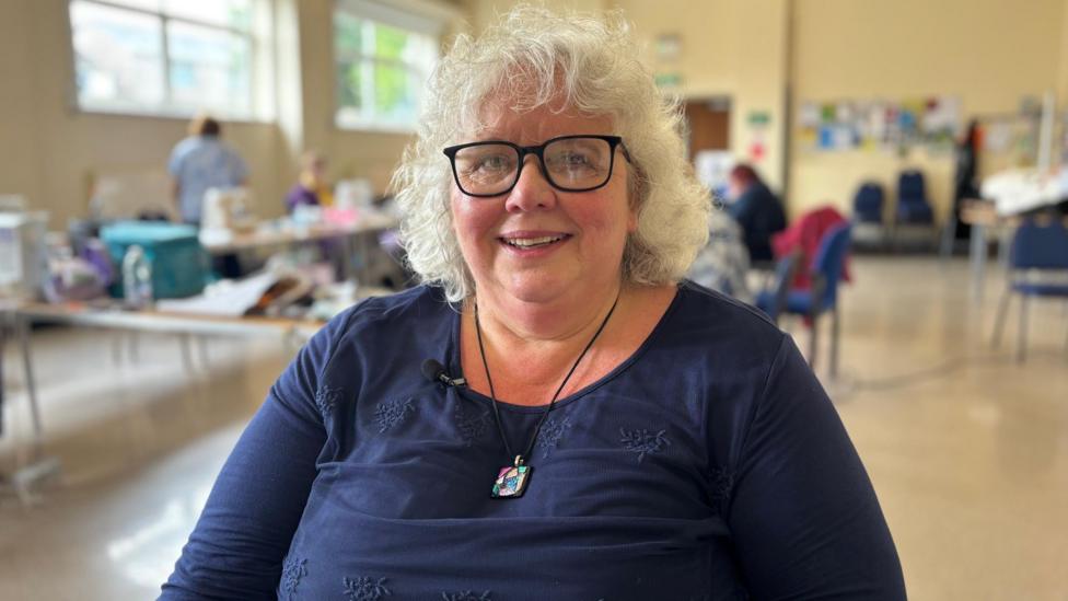Melinda Hawthorne is smiling. She has short, curly, white hair and black framed glasses. She is wearing a navy long sleeved top and a necklace with a colourful pendant.
