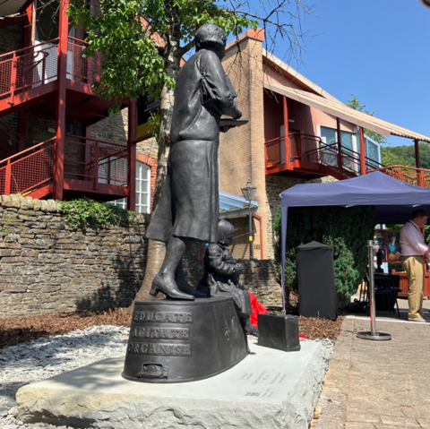 The back of a grey statue of a woman, who is wearing a skirt and heeled shoes. There is weiting on the plynth she is stood on which reads 