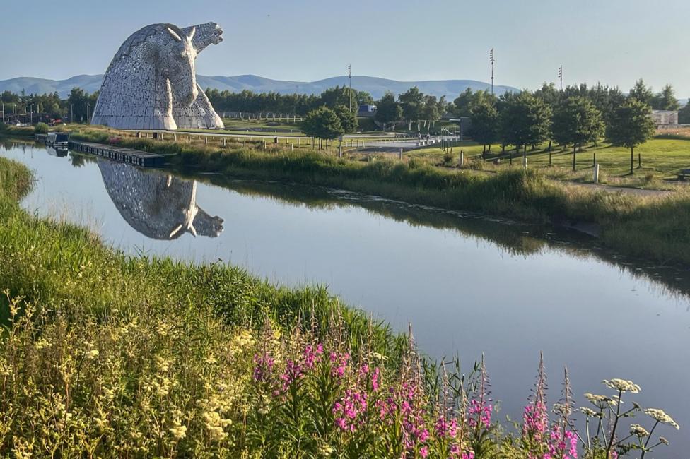 Large steel horse-head sculpture beside a calm canal, its reflection mirrored in the water, with green parkland, trees, and distant hills under a clear blue sky.