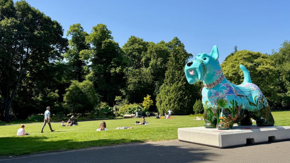 Colourful painted dog sculpture on a concrete plinth in a sunny park, with people relaxing on grass, trees behind, and a clear blue sky above.