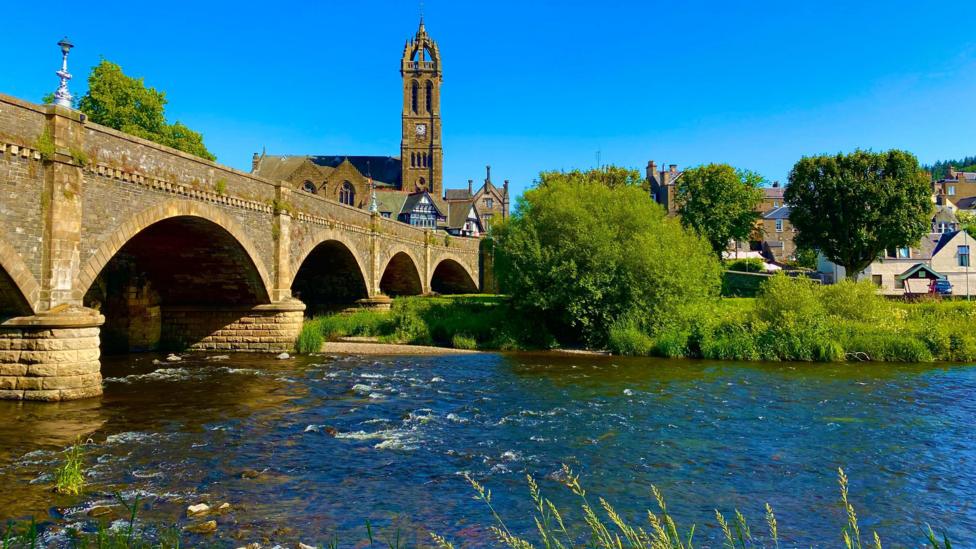 Historic stone arch bridge crossing a shallow river with flowing water, leading toward a town with buildings and a tall church tower under a bright blue sky, surrounded by green trees.