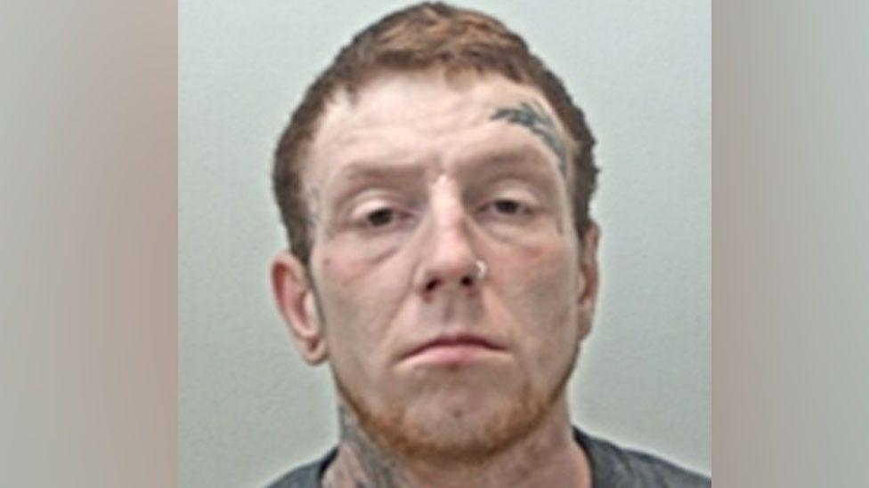 A police mugshot of Daniel Hardcastle. He has short red hair and a thin beard.