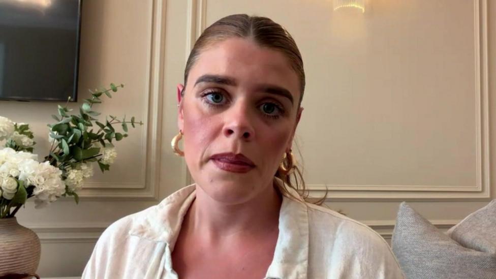 A woman with blonde hair scraped back into a pony-tail, red lipstick, gold earrings and a white linen shirt looks into the camera with a serious expression. 