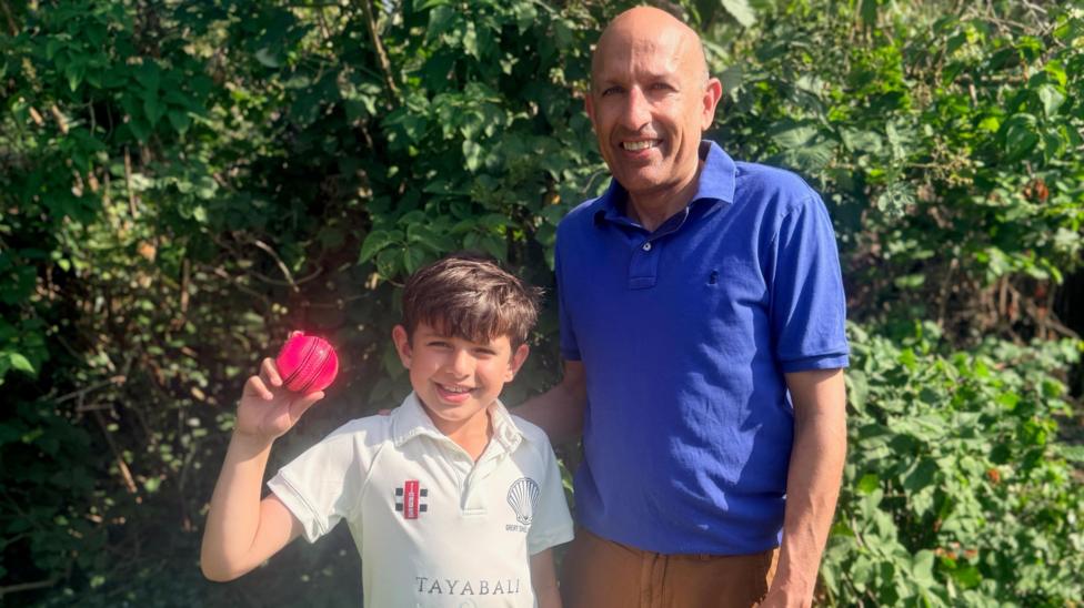 Rohan and Narain are in front of a hedge. Rohan is on the left and has short brown hair and is wearing a white cricket T-shirt. He is holding a red cricket ball near to his head. Narain is on the right and is wearing a blue polo shirt and orange trousers.