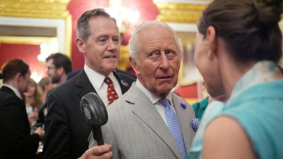 Vice Admiral Sir Tony Johnstone-Burt, Master of the Household to the Sovereign, wearing a black suit and red tie, holds a small battery powered fan for King Charles III, wearing a grey pinstripe suit and blue tie.