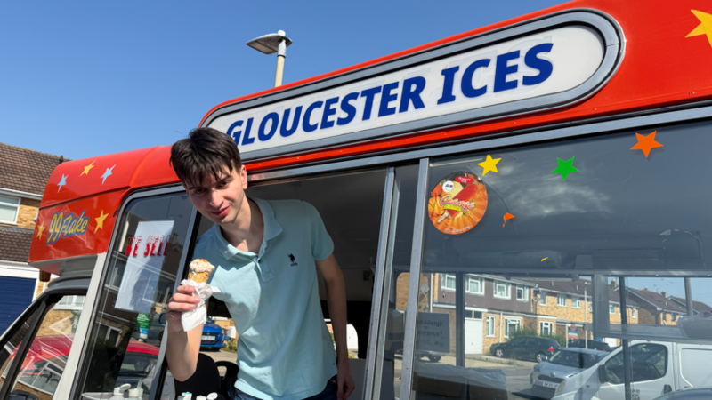 Emery, a young man in a blue polo neck shirt, smiles while holding an ice cream out of the window of an ice cream van, beneath a sign reading: 'Gloucester Ices'.