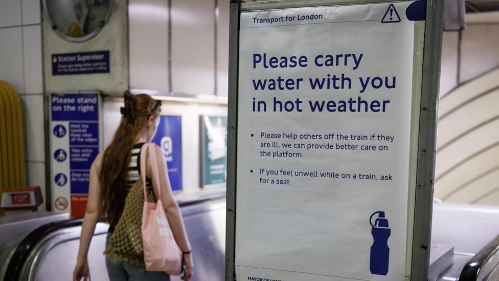 A woman with red hair carries a pink tote bag as she travels down an escalotor at a Tube station in London. Next to here is a TFL notice that reads 'please carry water with you in the hot weather'.