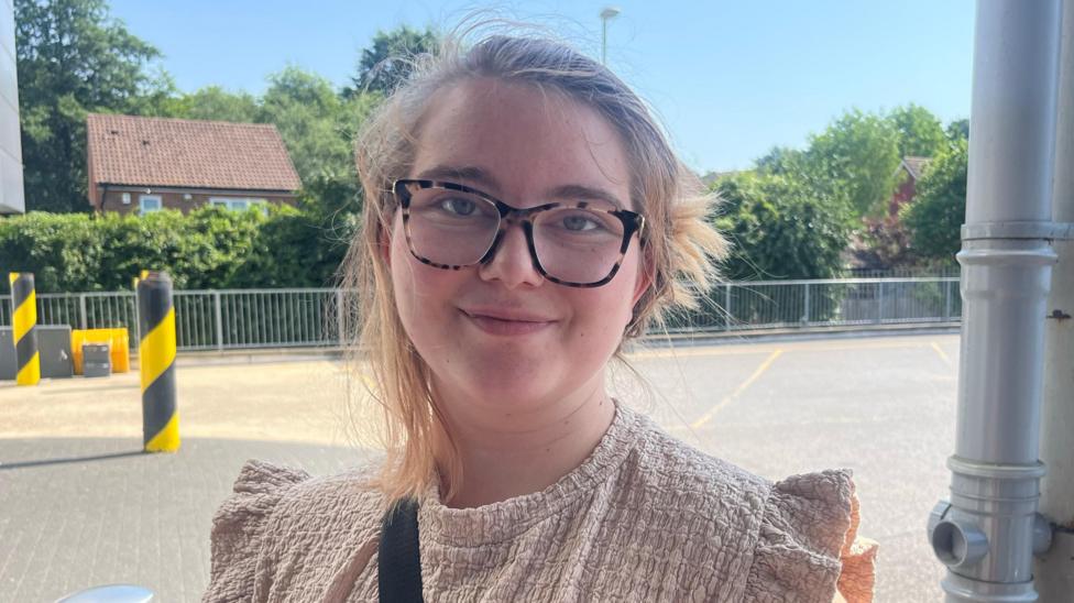 A young woman with tied-up blonde hair in a beige shirt. She is wearing black and brown glasses and there is a car park behind her.