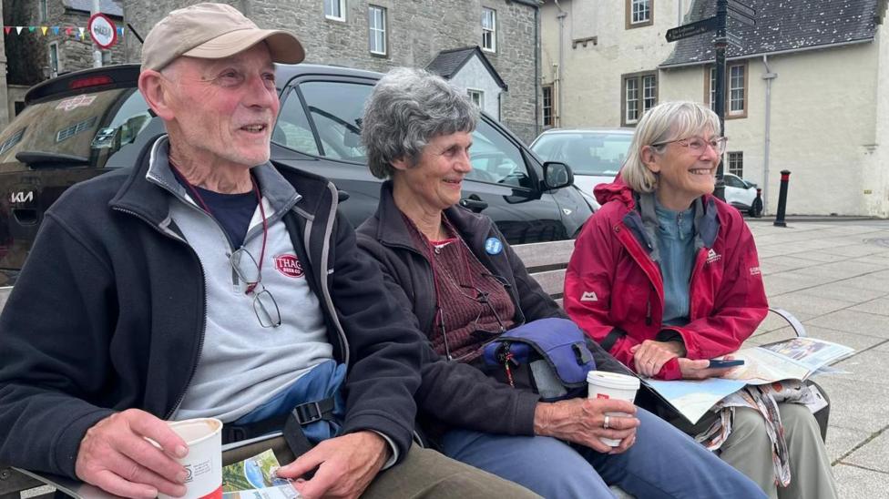 Ruth Nutman sits between two friends on a bench. Joy as short grey hair and she is wearing a black fleece over a burgundy top.