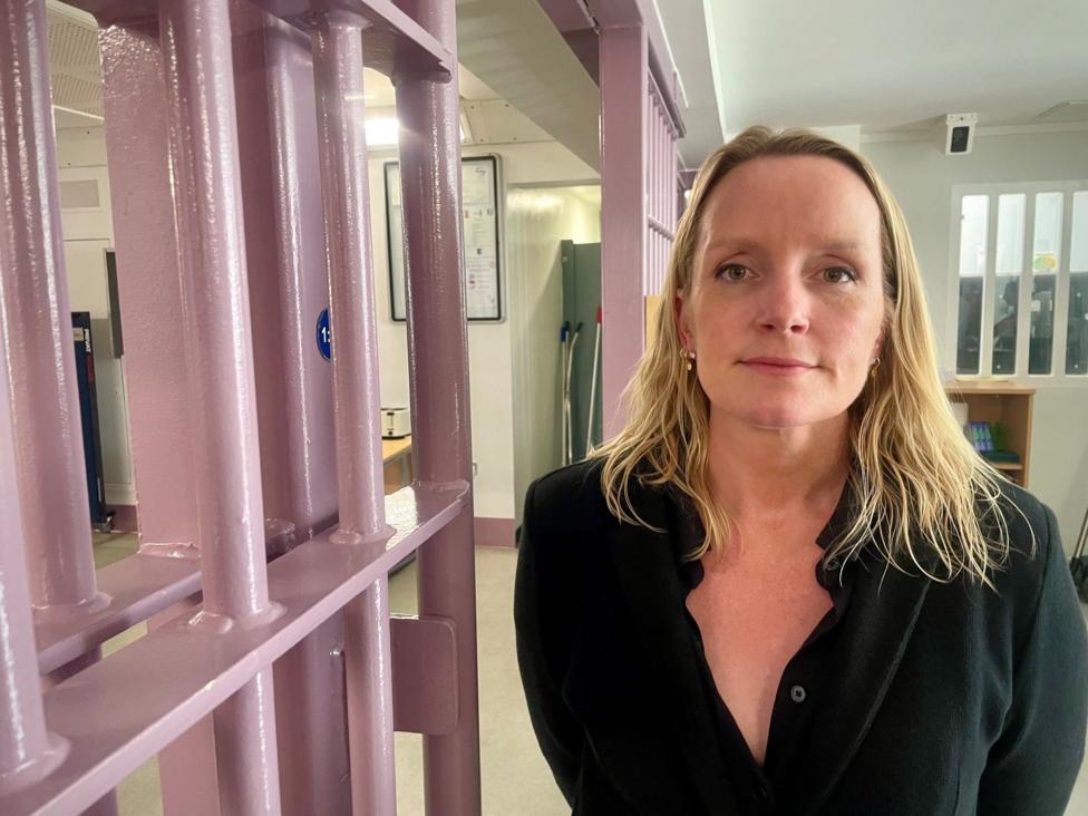 A woman with shoulder length blond hair, wearing a black shirt is looking at the camera. She is standing next to prison bars painted a light pink colour. 