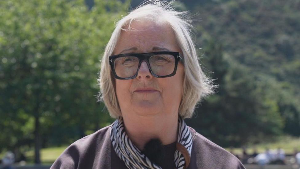 Cynthia Guthrie wears a brown top, zebra print scarf and dark glasses. She has short fair hair and is sitting outside. She looks slightly concerned 
