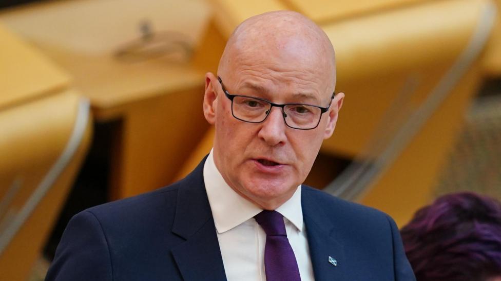 John Swinney wears dark glasses and wears a navy suit with a white shirt and purple tie in the debating chamber