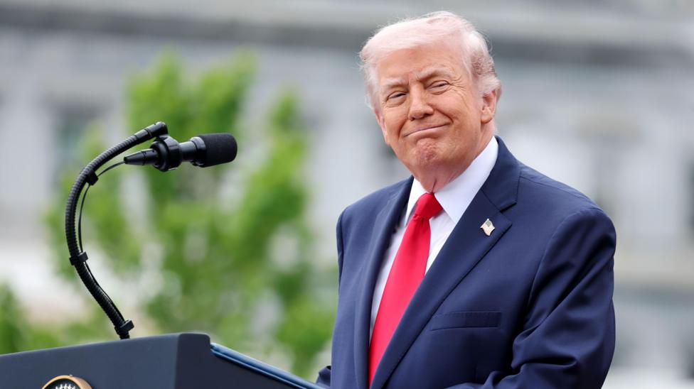 Trump standing at an outdoor podium with a microphone, wearing a dark suit and red tie, with blurred greenery in the background.