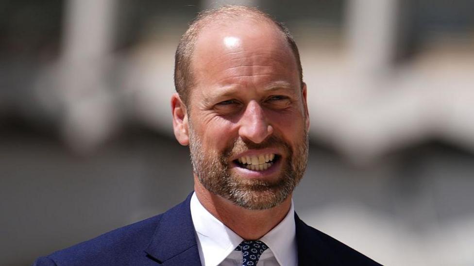 Prince William in a navy suit and white shirt smiles 