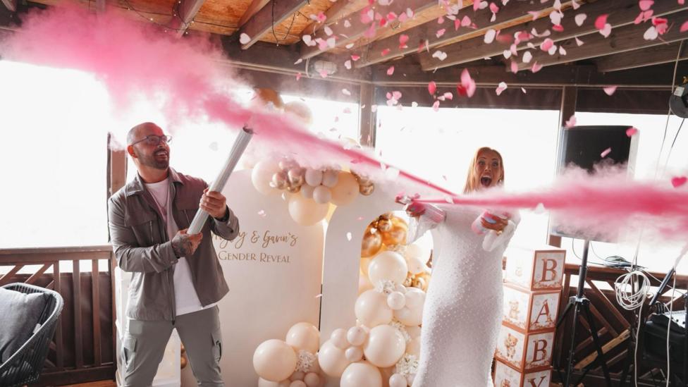 Hayley and her partner are each holing long white tubes with pink powder coming out. They are laughing, Hayley is dressed in all white. Her partner has very short hair and a beard and wears glasses. He is wearing grey trousers and a jacket over a white t-shirt, 