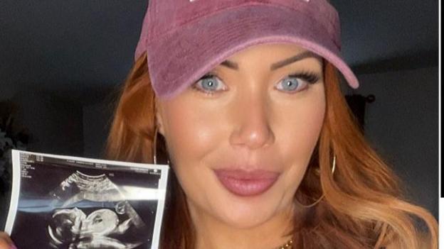 Hayley is holding her baby scan picture and smiling at the camera, She has long red wavy hair and is wearing a pink cap. 
