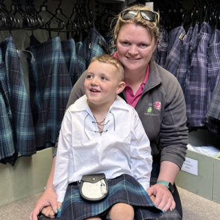 Gregor Corse is sitting on a small tartan stool with his mum Iona kneeling behind him. They are in a kilt shop. Gregor has short, blond hair and is wearing a kilt, sporran and traditional Scottish shirt with laces at the top. Iona has short, blonde hair and is wearing a pink T-shirt and a brown fleece. She has sunglasses on the top of her head and they are both smiling. There are a number of kilts behind them.