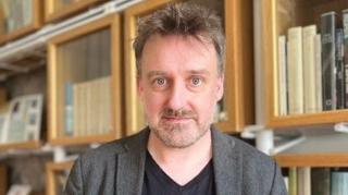 Petr Mackay is looking at the camera with shelves full f books behind him. He has short, grey/brown hair and is wearing a black T-shirt and a grey suit jacket.