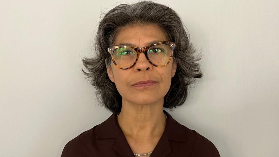 Close-up portrait of Sarah Armstrong wearing round tortoiseshell glasses and a dark collared top, facing the camera against a plain light-colored background.