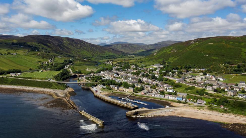 An aerial view of the village of Helmsdale on the east Sutherland coast. The village has a culster of houses and other buildings on a hillside, and there is a historic harbour. Green hills rise behind the village. It is pictured on a sunny day with white, fluffy clouds in the sky.