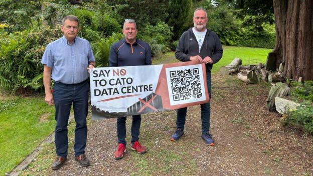 Three men stand on a garden path surrounded by green plants with a banner that reads say no to the Cato Data Centre