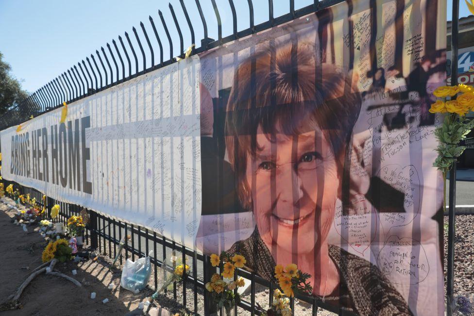 A long banner is attached to some black railings, with flowers also laid at its base. The words 'BRING HER HOME' are emblazoned on the banner with the face of Nancy Guthrie on the right hand side