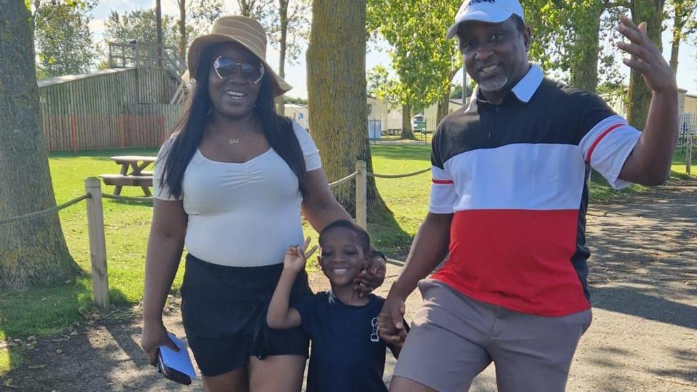 A man and woman and young son hold hands and smile in the sunshine. They are all dressed in summer clothes