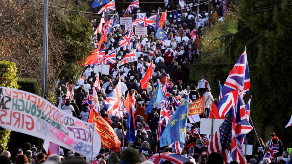 A large group of protestors. They are holding Union Jack and local flags. 