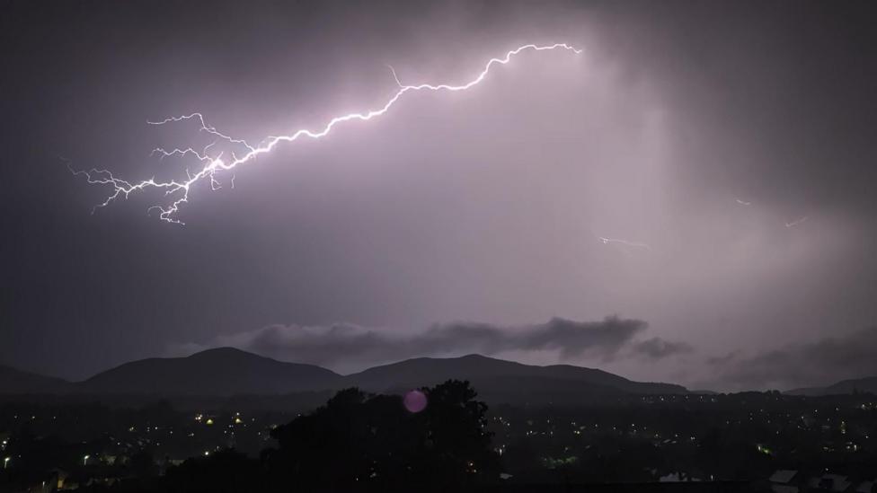 A dark night sky with the a bright lightning bolt. There are dark hills in the background.