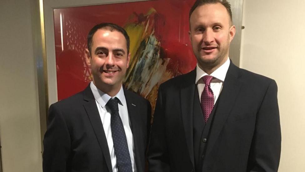 Two men with short brown hair are wearing a shirt and tie and smiling at the camera. 