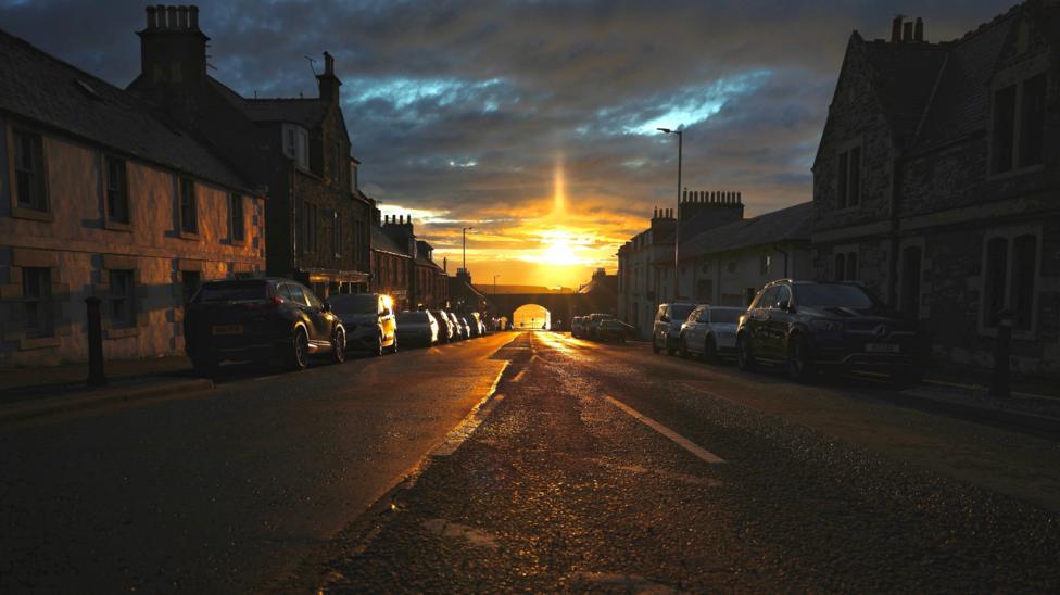 A dark village street with cars parked along the pavements. There is bridge with an archway at the end of the road. The sunset reflects orange along the street and the sky is dark.