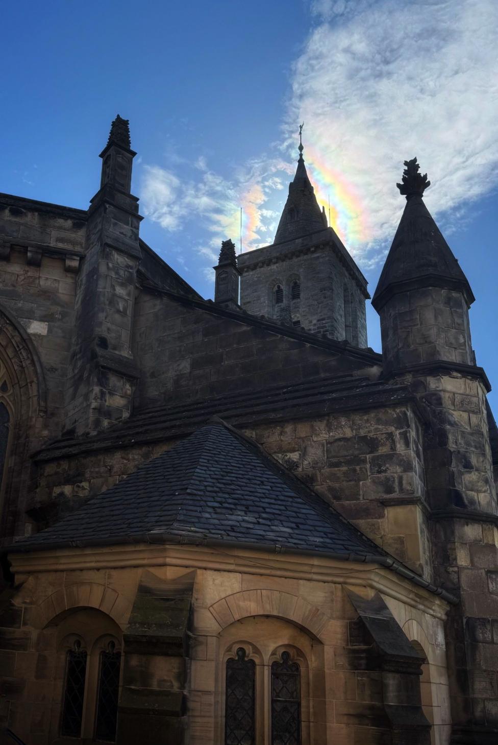 A large chapel with turrets and spires. A rainbow iridescence cloud is above the tower. There are white clouds on the left and the sky is clear blue.