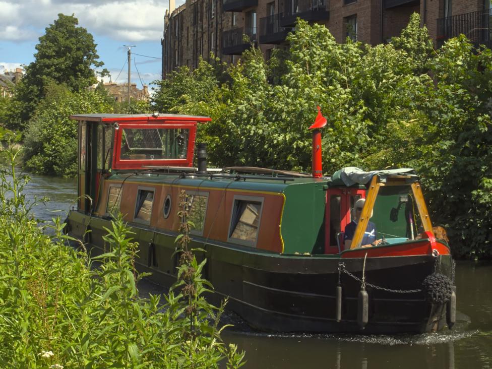 A red and orange canal boat in the water. Trees and bushes line the canal on both sides.