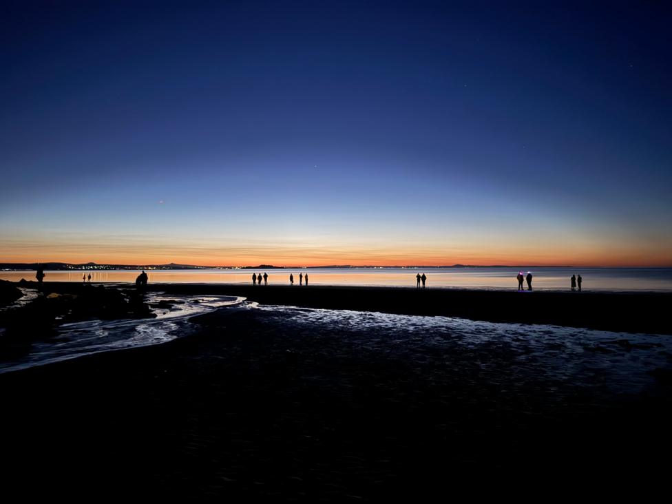 A dark beach. There are people standing at the water's edge and the horizon is orange.