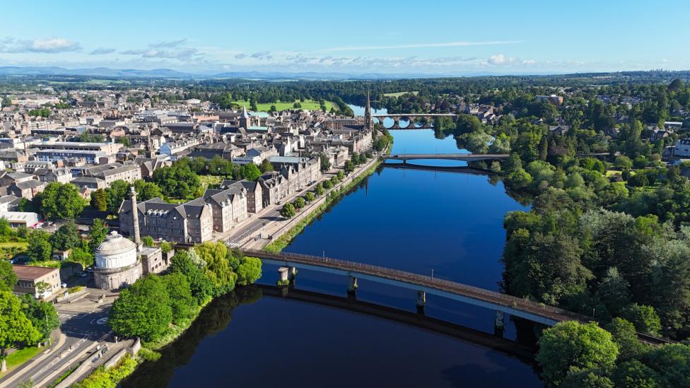 A birdseye view of Perth. The River Tay runs through the middle of the city and there are three bridges crossing it. Buildings and trees line either side of the river.