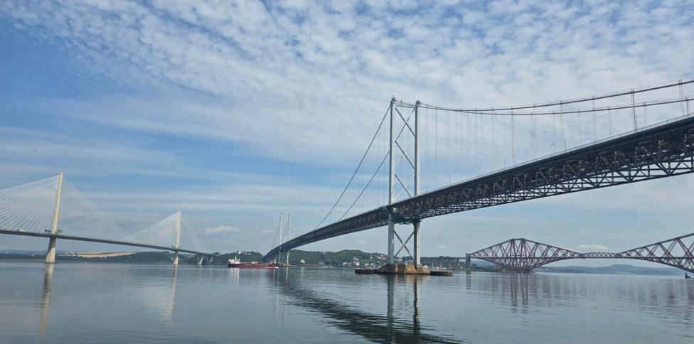 The Queensferry Crossing, Forth Road Bridge and Forth Rail Bridge spanning across the water. The reflections of the bridges are in the water and there are clouds in the sky.