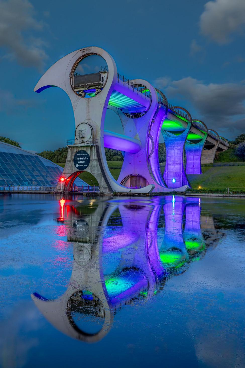 The Falkirk Wheel illuminated in purple, green and red at night. The reflection is in the water and the sky is dark with clouds.