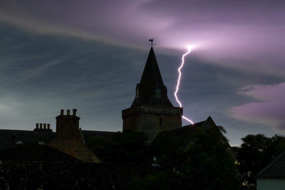 Dornoch Cathedral under the night sky. It is dark and there is a lightning bolt in the purple-coloured sky.