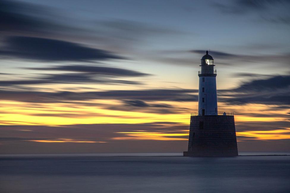A lighthouse in the sea. The sky is orange with dark clouds streaked across it.