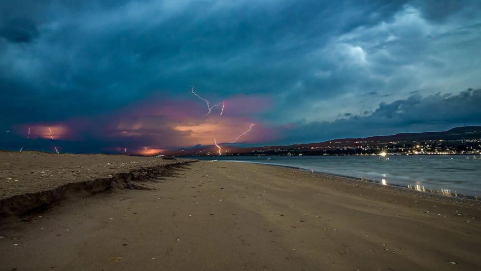Shows a derelict beach with water to the right and dark skies lit up by lightning 