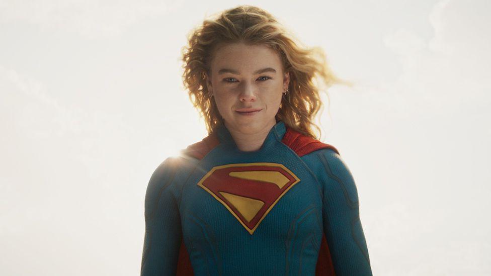Milly Alcock, wearing the blue Supergirl suit complete with red cape and red S emblazoned on the chest, stands against a hazy, sunny sky and stares down the camera with a wry smile on her face.