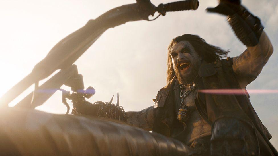 A screengrab from Supergirl shows Jason Momoa as Lobo, dressed in a leather vest, riding a motorbike and roaring as he speeds towards his destination.