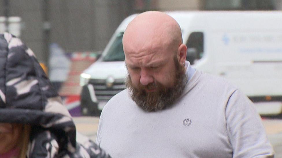 Alan Keelan arriving at court wearing a light grey t shirt. He has a bald head and a light brown beard.