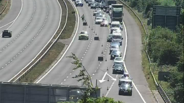 Motorway camera image of cars being sent off the motorway. There are long queues as cars and lorries make their way off the motorway with police and cones in view.