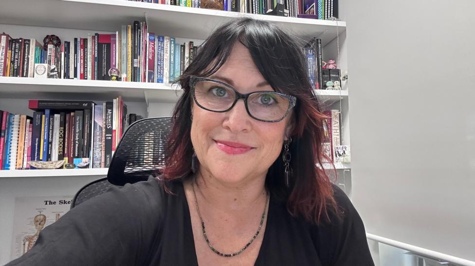 Dr Trish is sitting in front of a bookcase in a white office. She is wearing a black top, glasses, a necklace and has black hair with reddish/pinkish tips. She is wearing pink lipstick and smiling at the camera. 