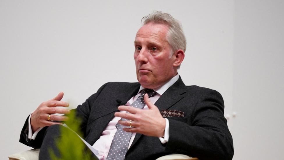 A man in a dark suit and light pink shirt and tie is sitting against a plain white wall, looking to the left of the camera. He has grey hair and a hankerchief in his breast pocket. His hands are open and in the air mid-movement while he speaks. 