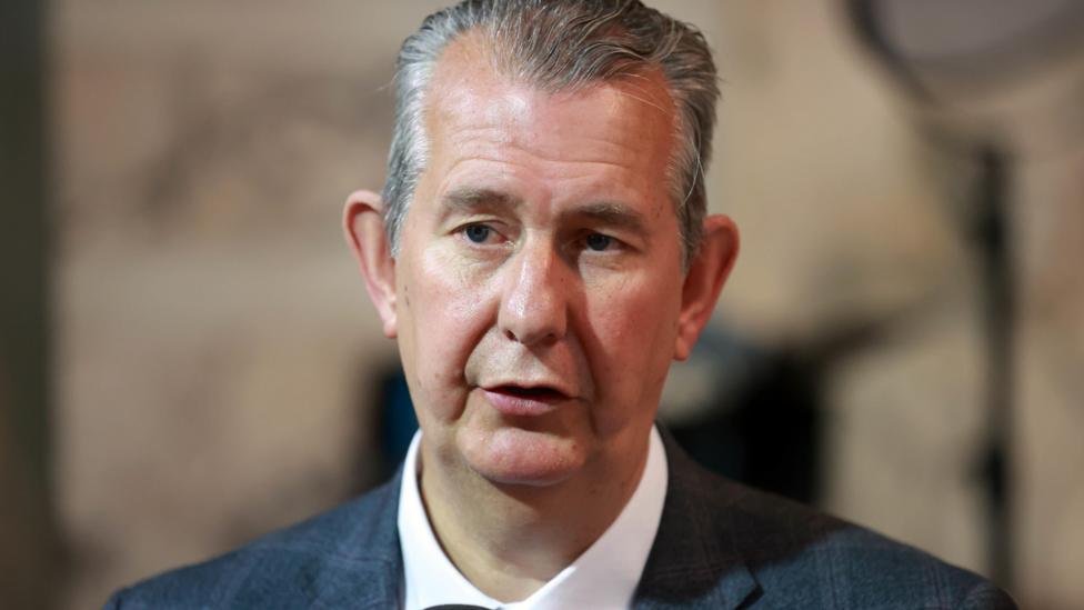 A man with dark, greying hair, is looking to the left of the camera He is wearing a suit with a white shirt and is only visible from the shoulders up. Behind him is a blurred background. 