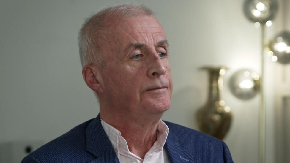 A man with short grey hair looks to the right of the camera. He is wearing an open neck shirt and navy blue blazer Behind him is a tall lamp, illuminating the dimly lit room. 
