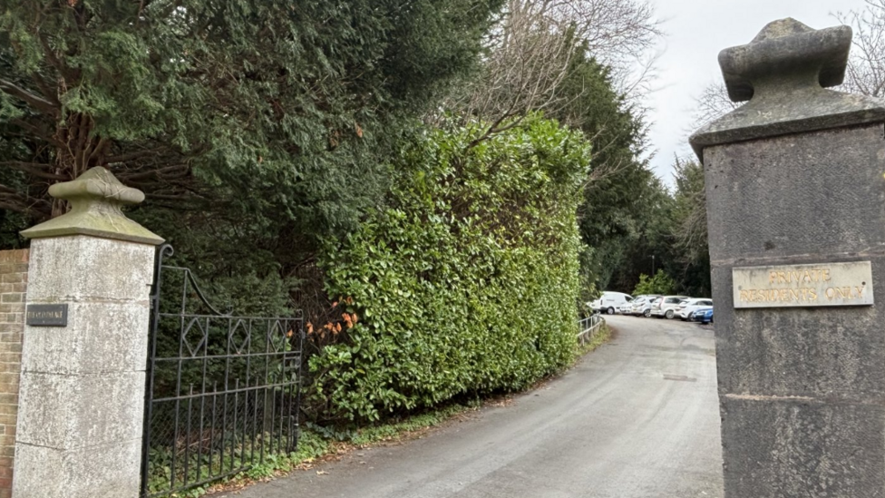 Two pillars on either side of a long driveway with a green hedge along one side and a black gate swung open.